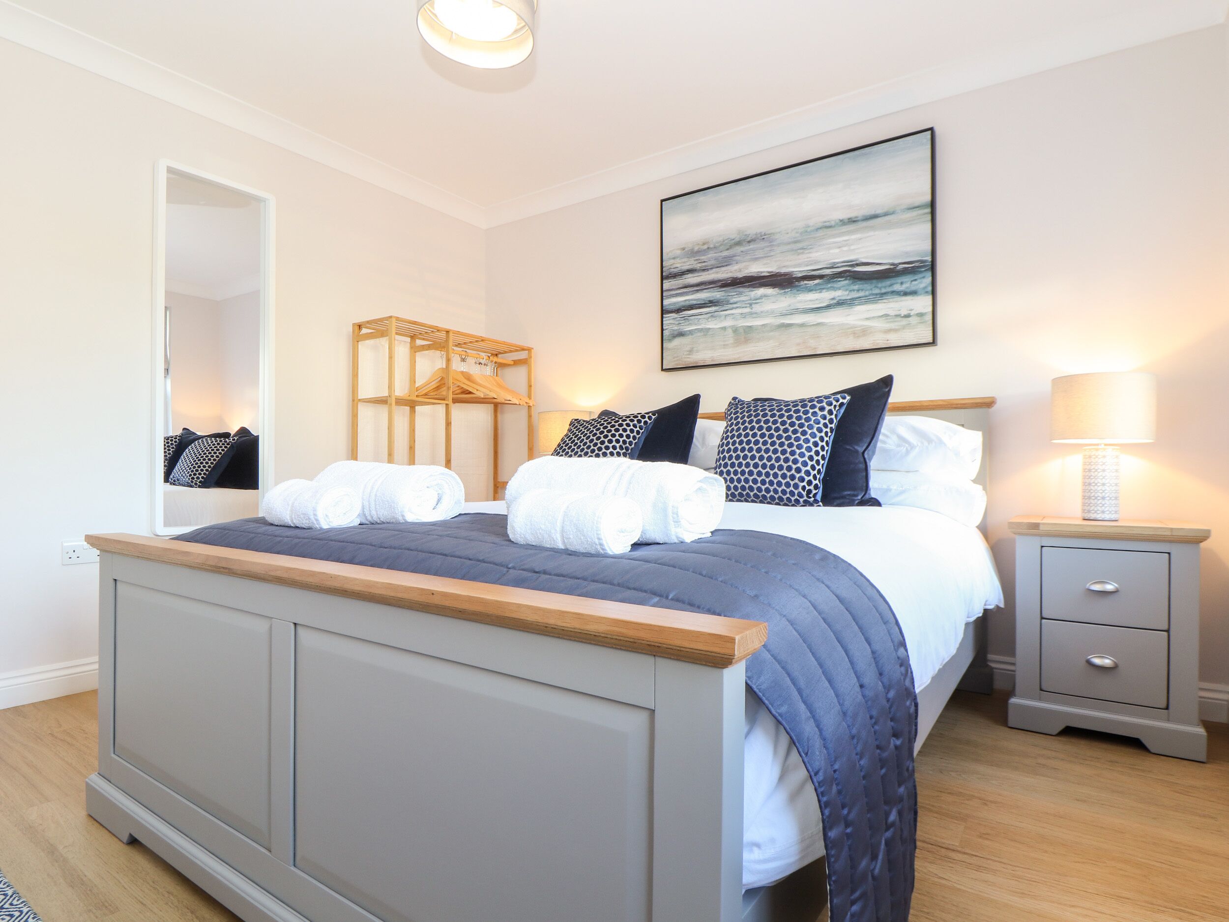 A modern bedroom with a large bed, a bedside table, a nightstand, a mirror, and a painting on the wall.