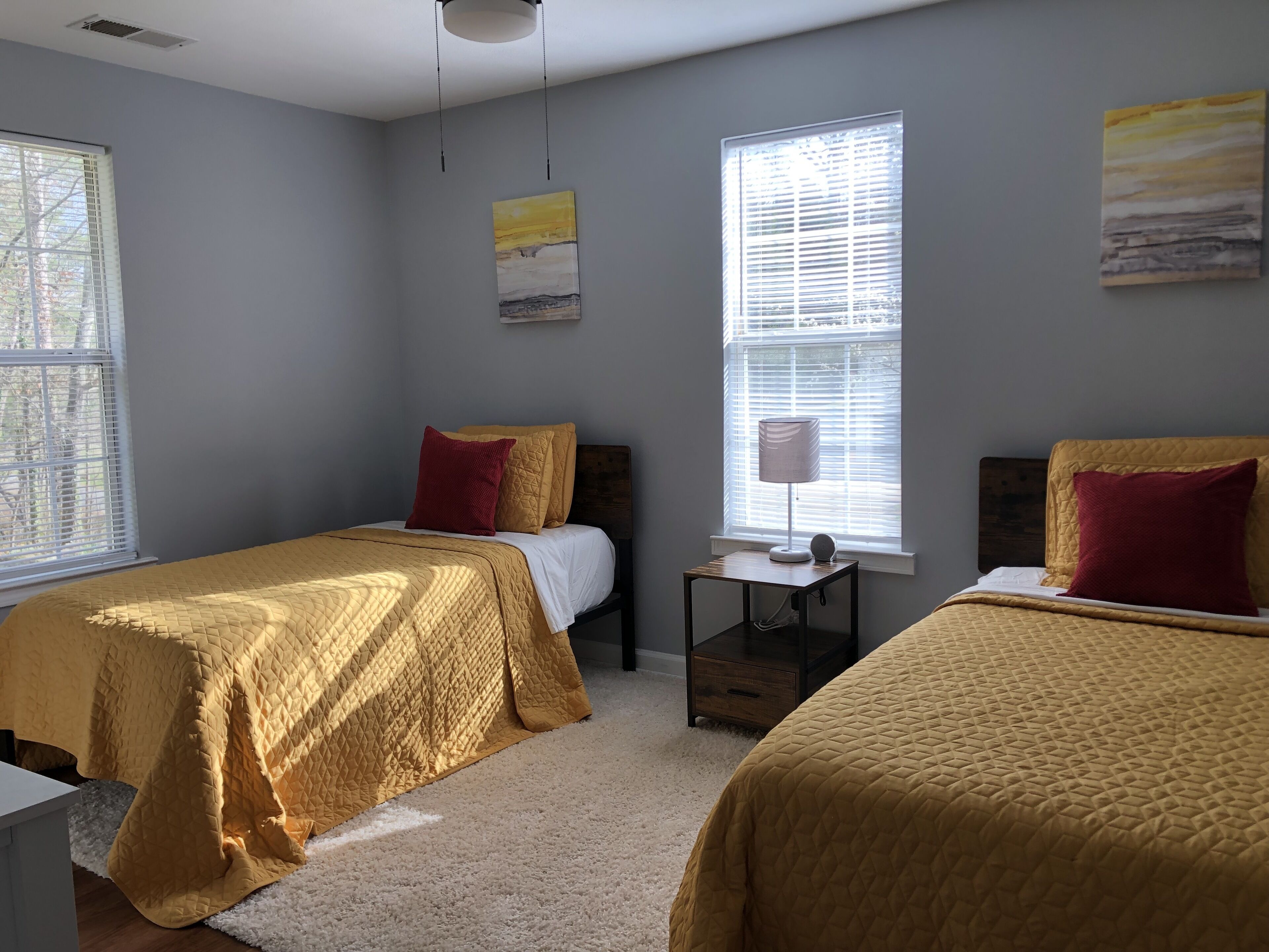 A bedroom with two beds, each with a yellow quilted bedspread and red pillows. There is a bedside table with a lamp and a small decorative item. The room has a window with blinds, a ceiling light, and artwork on the walls.