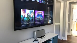 A flat-screen TV mounted on a wall displaying video game titles and a PlayStation console on a white stand. - Cozy Farmhouse near Atlanta's Airport! (Atlanta)