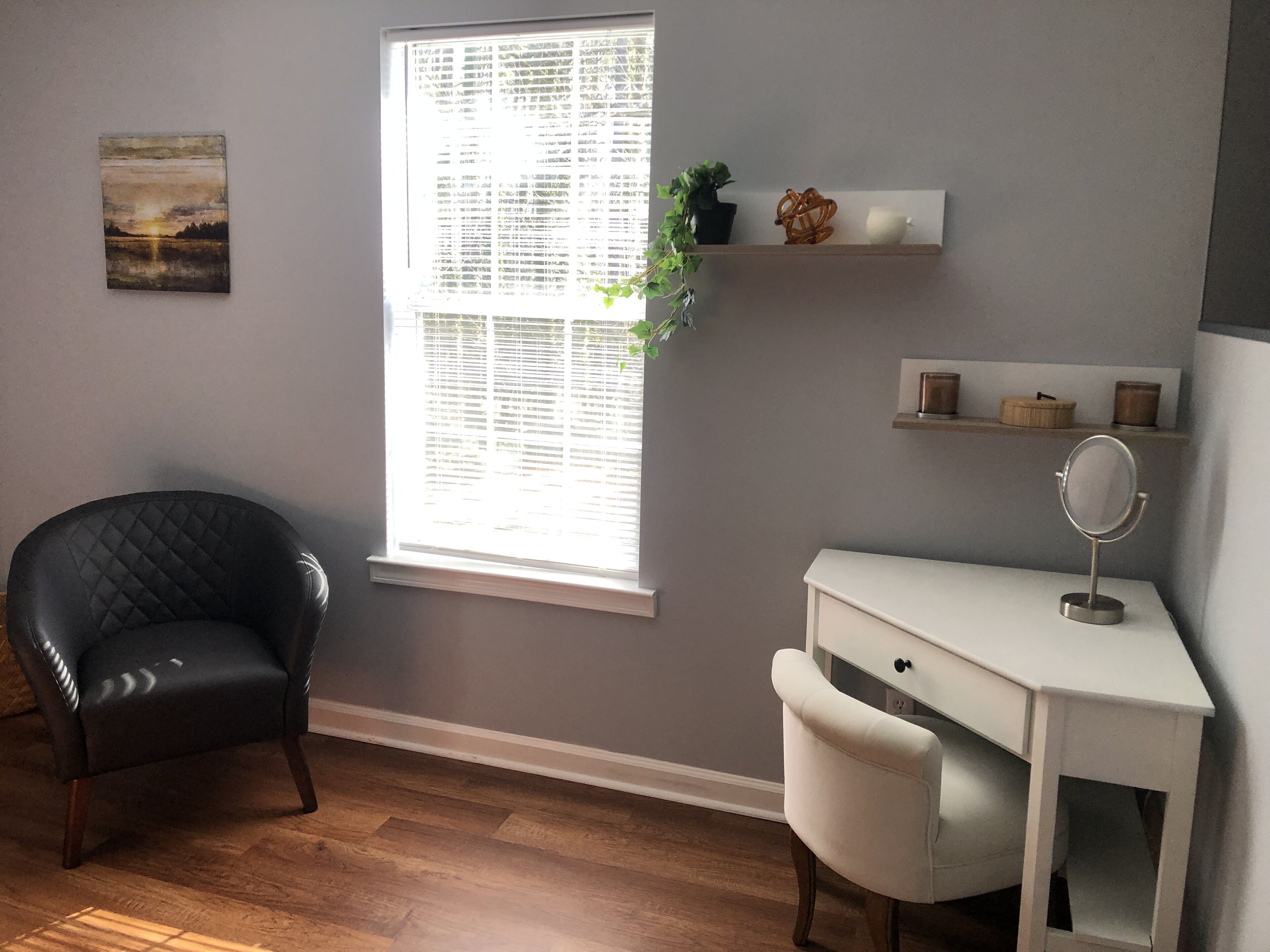 Une pièce avec un fauteuil en cuir noir, un bureau blanc avec un miroir et une fenêtre avec des stores.