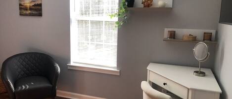 Une pièce avec un fauteuil en cuir noir, un bureau blanc avec un miroir et une fenêtre avec des stores.