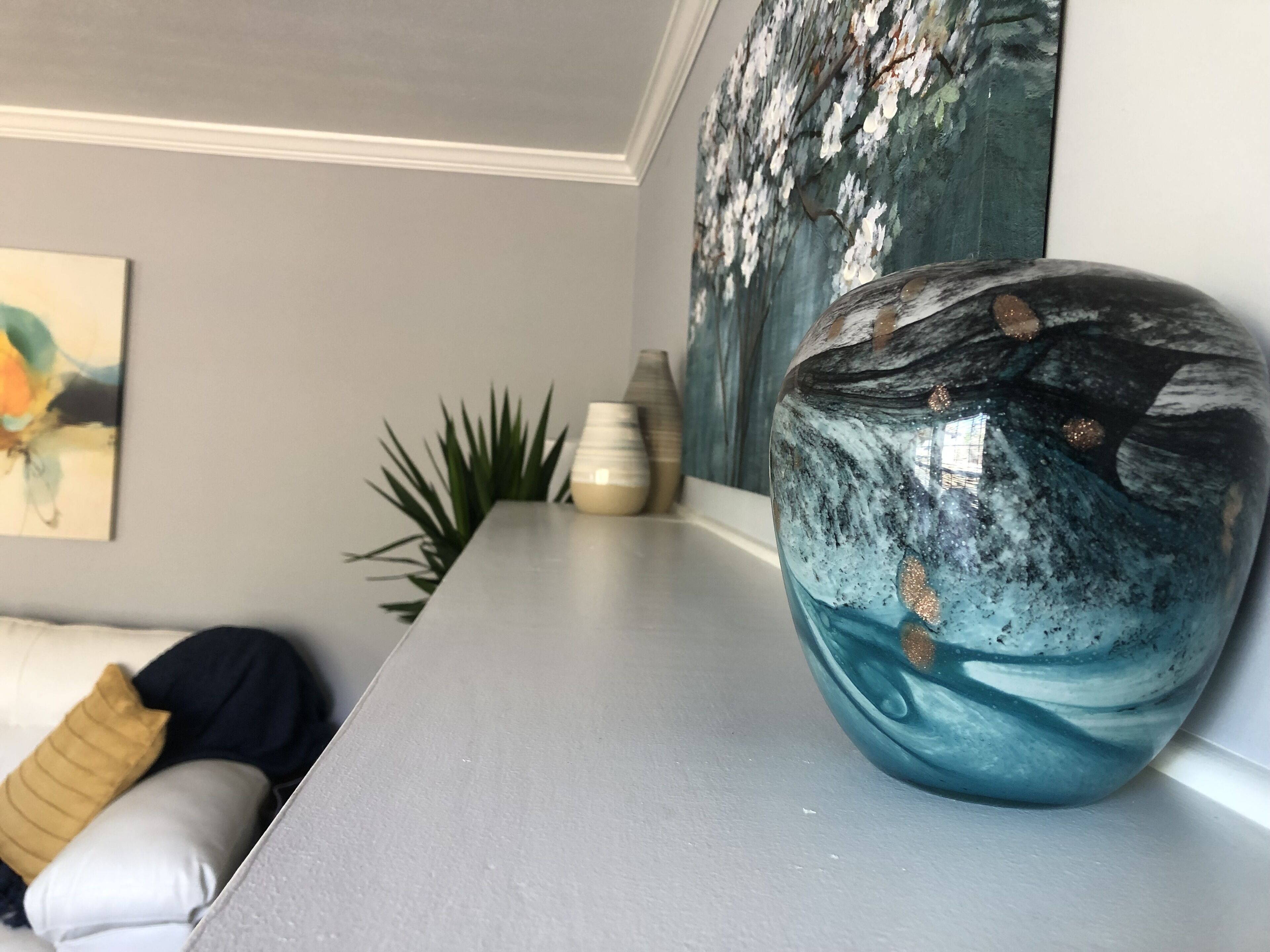 A modern living room with a large decorative vase on a grey table, a painting on the wall, and a plant in the corner.