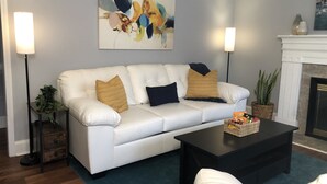 A living room with a white leather sofa, two floor lamps, a fireplace, and a painting on the wall. - Cozy Farmhouse near Atlanta's Airport! (Atlanta)