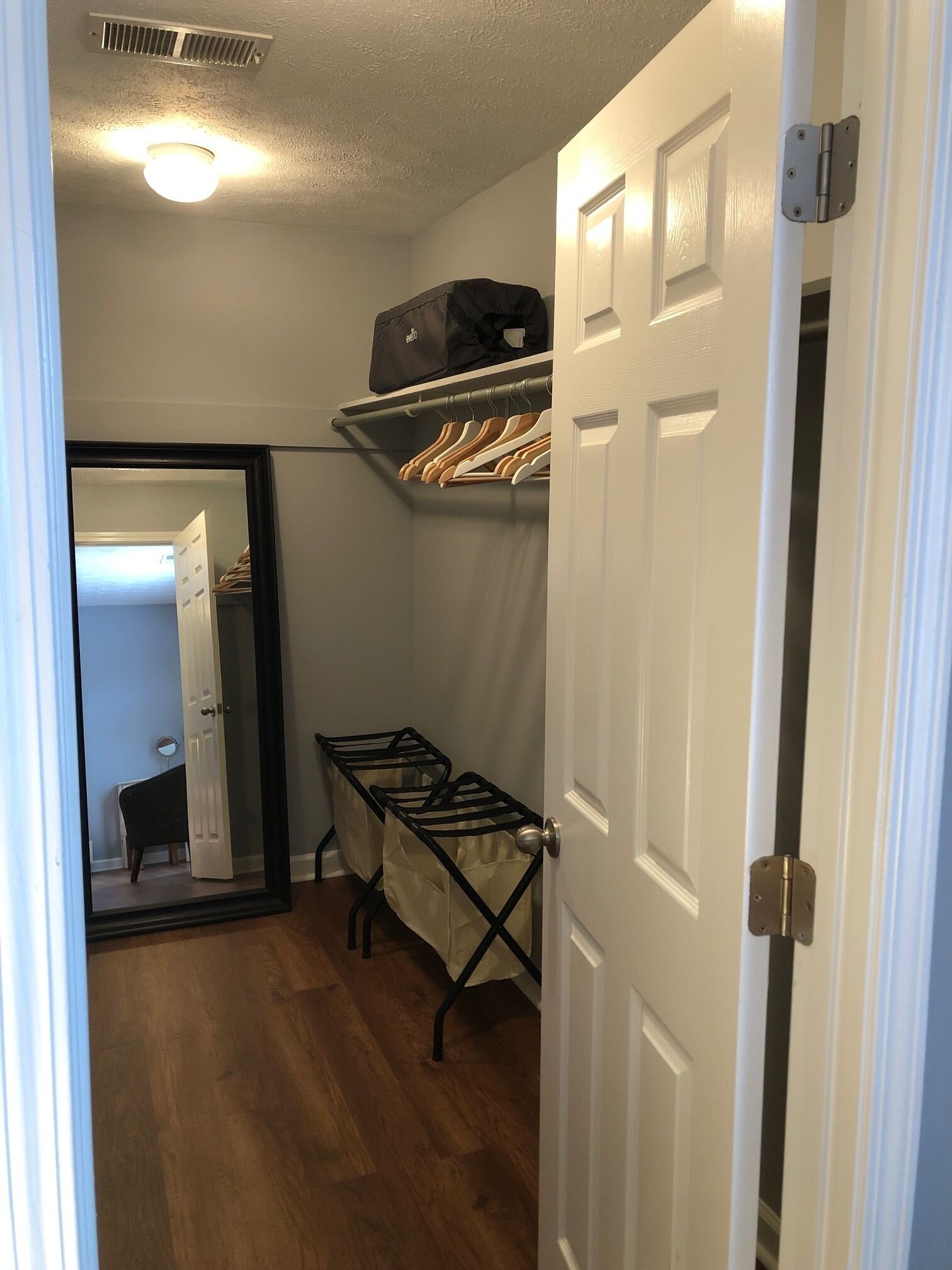 A narrow hallway with a wooden floor, a ceiling vent, a hanging clothes rack, and a door with a mirror.
