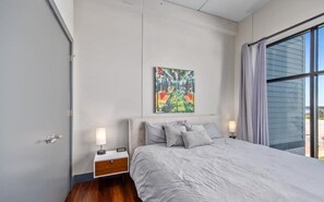 Une chambre à coucher moderne avec un grand lit, une table de chevet, un tableau au mur et une fenêtre avec des rideaux.