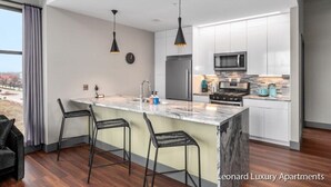 A modern kitchen with white cabinets, a marble island, black pendant lights, and a stainless steel refrigerator. - Spacious Luxury Apartment with Waterfront Views - The Leonard Building (Muskegon)