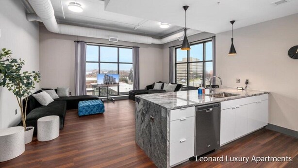A modern apartment with a kitchen island, a dining area, and a living room with a sofa and a blue ottoman. - Spacious Luxury Apartment with Waterfront Views - The Leonard Building (Muskegon)