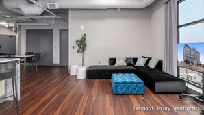 A modern apartment with a large black sectional sofa, a small blue ottoman, a dining area with a white table and chairs, and a view of the city through large windows. - Spacious Luxury Apartment with Waterfront Views - The Leonard Building (Muskegon)