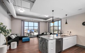 A modern apartment with a kitchen island, a dining area, and a living room with a sofa and a blue ottoman. - Spacious Luxury Apartment with Waterfront Views - The Leonard Building (Muskegon)