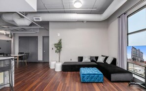 Een modern appartement met een grote zwarte modulaire bank, een klein blauw voetenbankje, een eethoek met een witte tafel en stoelen, en een stadszicht door grote ramen.