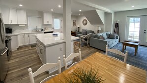 A modern kitchen with white cabinets, a central island, and a dining area with a wooden table and chairs. - Treasured Memories* Steps to sand * Exclusive Patio * REMODELED * CLEAN (Newport Beach)