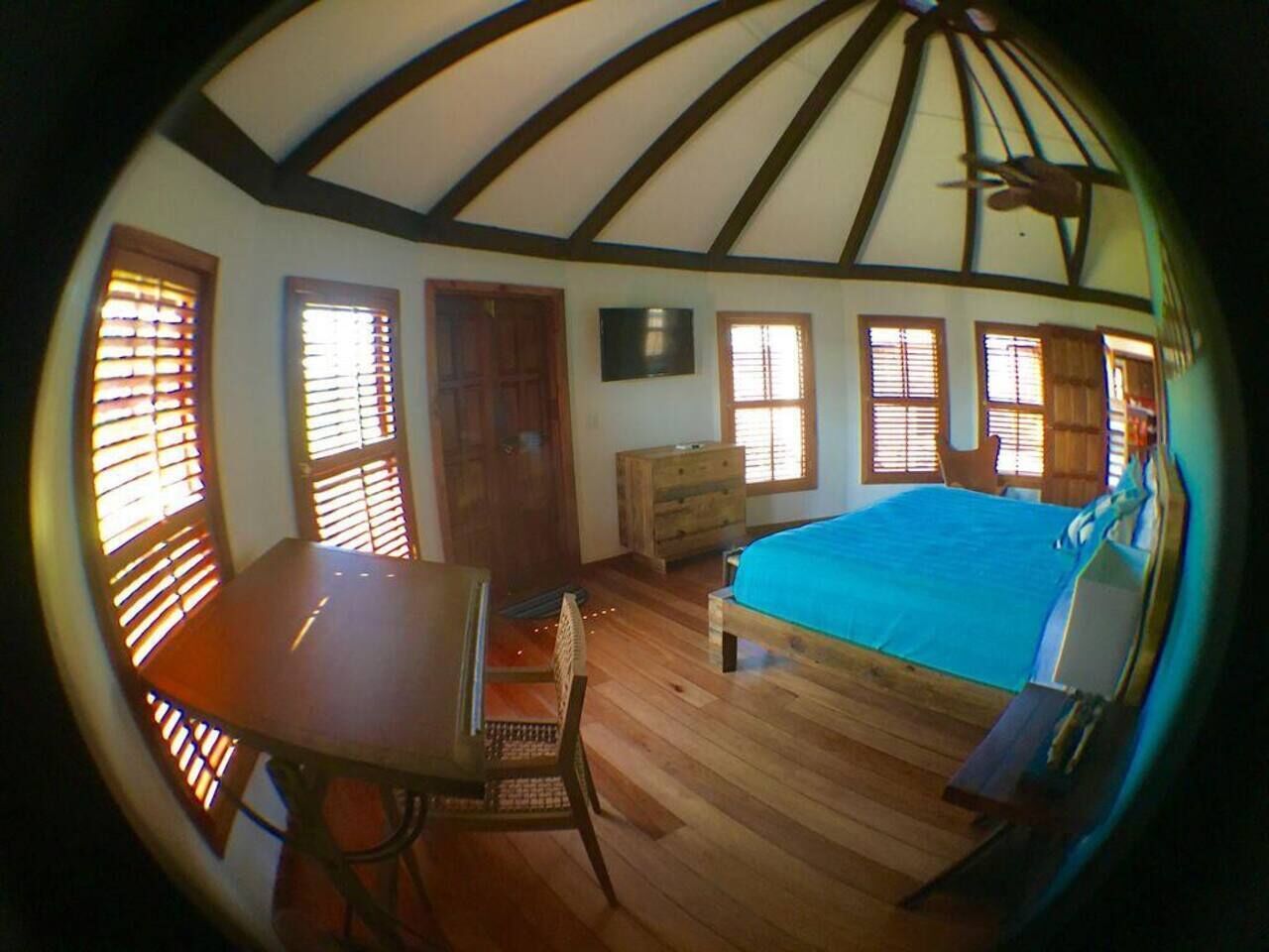 Ein Zimmer mit einem Bett, einem Tisch und einem Stuhl. Es hat einen Holzboden und eine Decke mit einem Ventilator.