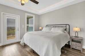 A bedroom with a bed, two bedside lamps, a nightstand, a ceiling fan, and a door leading to a balcony. - Fishermans Delight 3 Bedroom Home by RedAwning (Port Bolivar)
