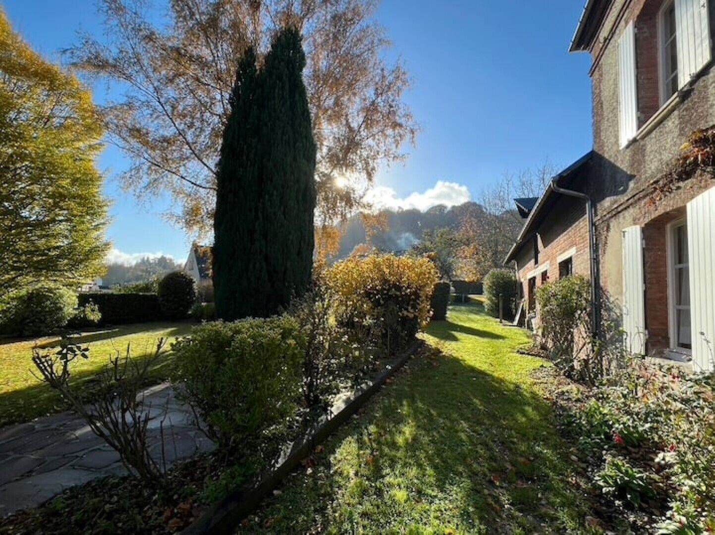 Une propriété résidentielle dotée d’un jardin bien entretenu, d’un grand conifère et d’une maison en briques avec des volets blancs.