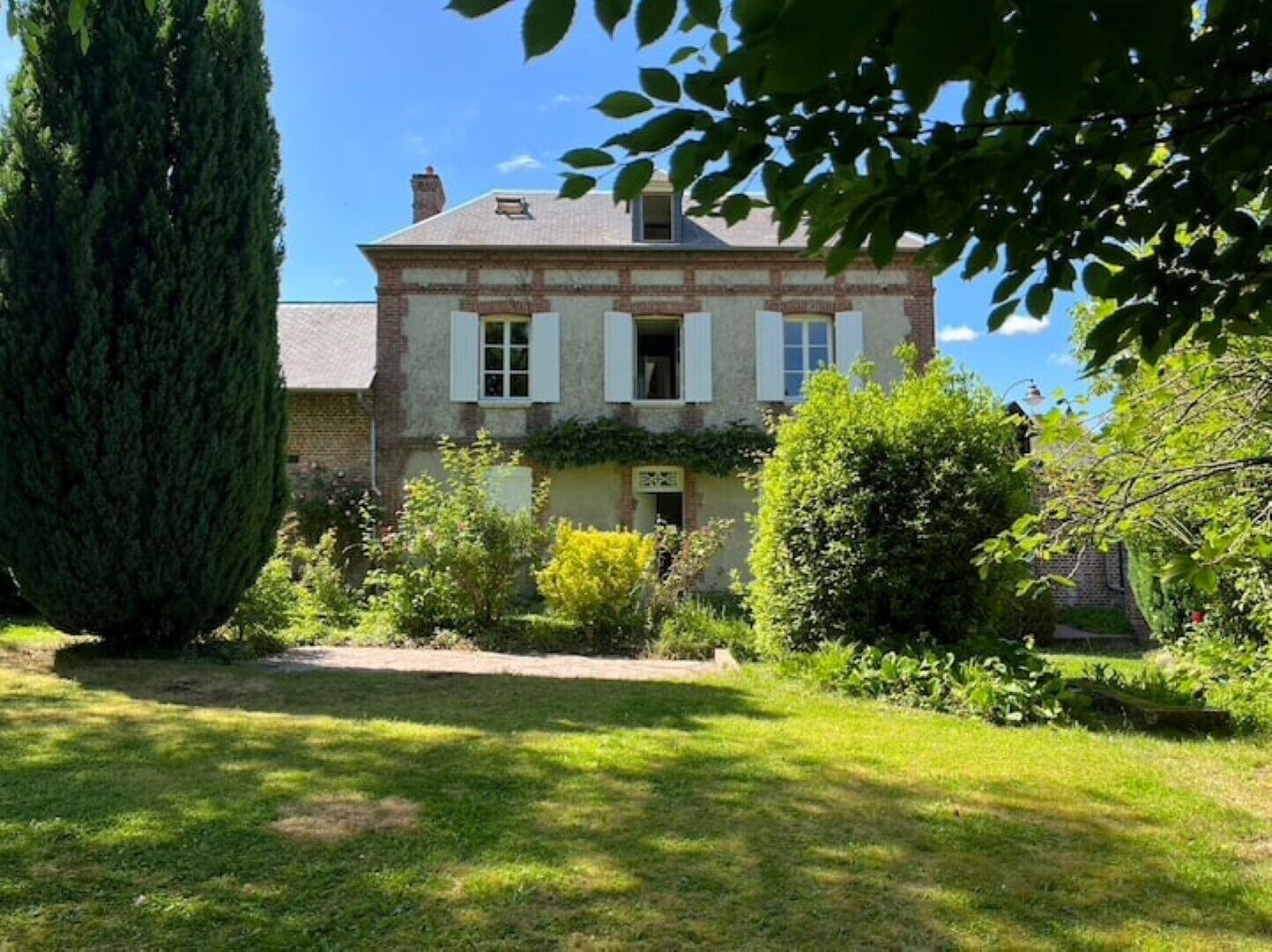 A traditional two-story house with a well-maintained garden, a tall tree, and a clear blue sky.