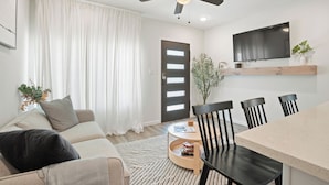 Un salon moderne comprenant un canapé, une table basse, une table à manger avec des chaises, un ventilateur de plafond, un téléviseur fixé au mur et une porte avec une fenêtre.
