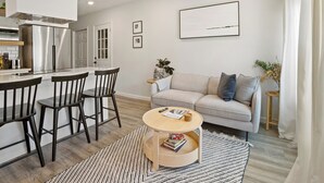 Une cuisine moderne dotée d’appareils électroménagers en acier inoxydable, un coin repas avec des chaises foncées, une table ronde en bois et un canapé de couleur claire.