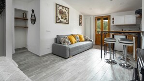 A modern living room with a grey sofa, wooden shelves, a kitchen with a wooden door, and a dining area with bar stools.