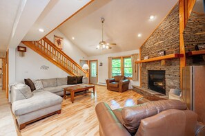 A spacious living room with a stone fireplace, wooden staircase, and a large window.