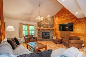 A cozy cabin interior with a stone fireplace, wooden walls, a ceiling fan, and a flat-screen TV.