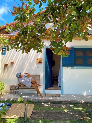 A person relaxing on a wooden bench outside a house with a blue door and white walls. - Pousada Céu de Corumbau (Prado)