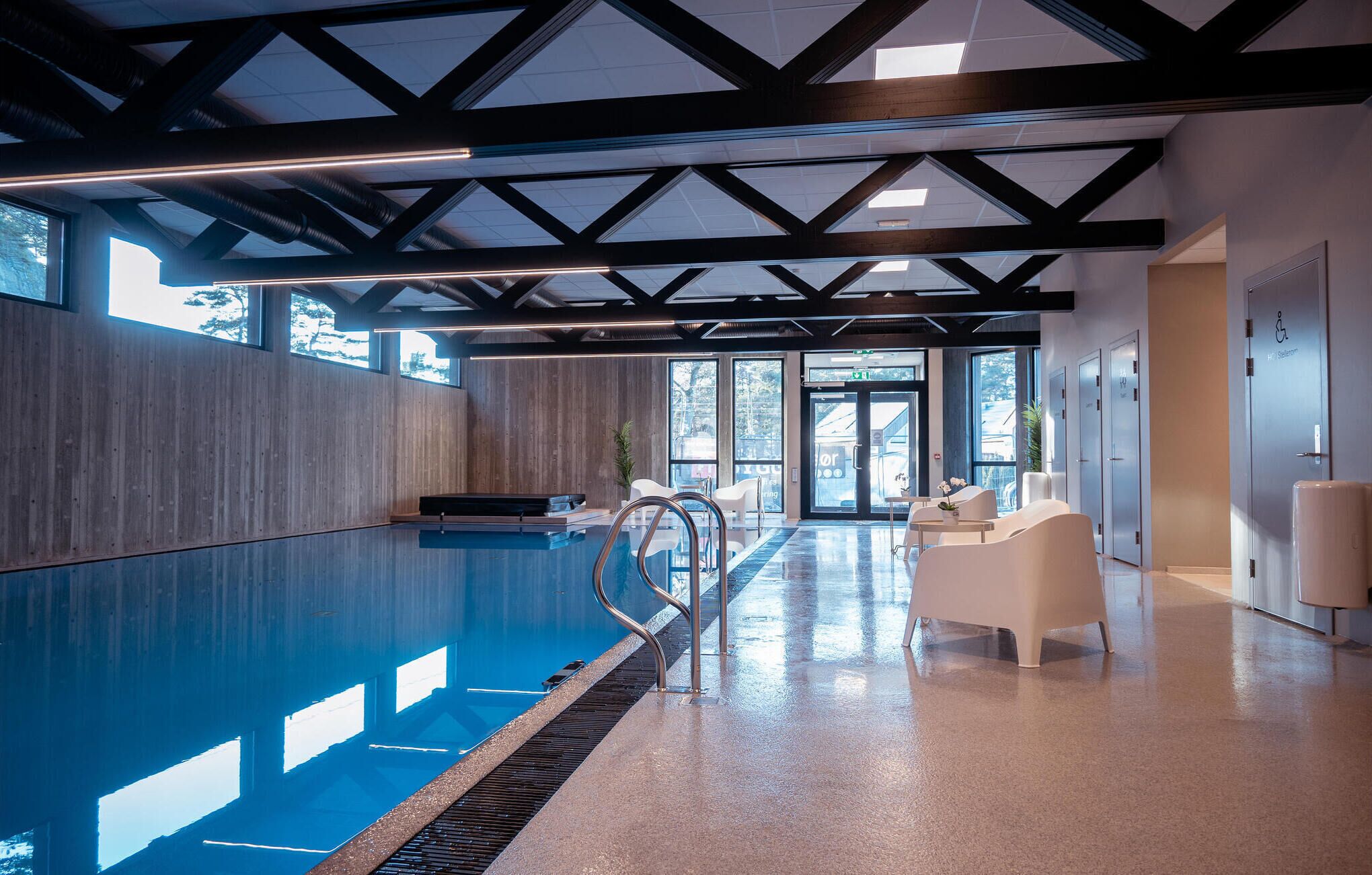 Ein modernes Hallenbad mit freiem Blick zum Himmel durch große Fenster, einer Metallleiter und einer Sitzgelegenheit mit weißen Stühlen.