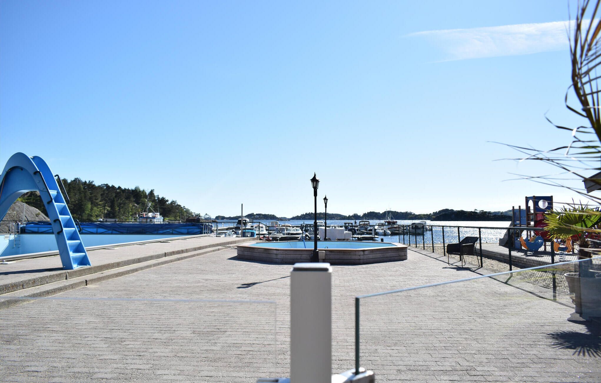 A waterfront promenade with a blue pedestrian bridge, a circular seating area, and boats docked in the background.