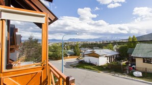 Un balcón de madera con vistas a una zona residencial, una calle y montañas al fondo.