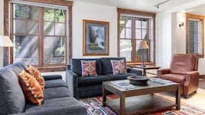 A cozy living room with a dark gray sofa, a brown leather armchair, and a wooden coffee table. There are large windows with wooden frames, a framed picture on the wall, and a lamp on a side table. - Outpost: Grand View 5020A - Fireplace, Elevator, AC! (Jackson)
