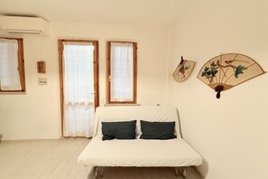 A room with a bed, two windows with curtains, a wall-mounted fan, and a small picture frame.