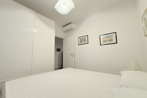 A bedroom with a bed, white walls, two framed pictures, an air conditioning unit, and a door leading to another room. - M333 - Marcelli, cozy two-room apartment 200 meters from the beach (Marcelli)