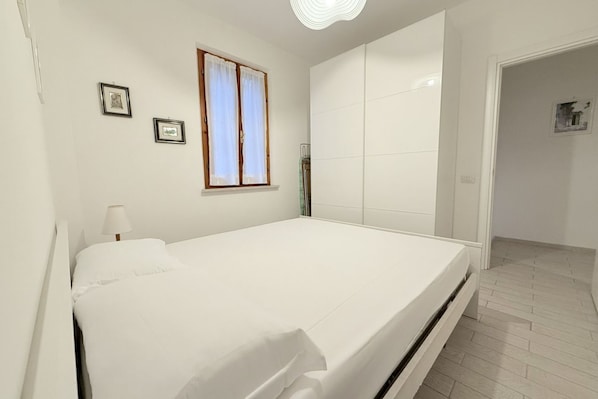 A bedroom with a bed, a window with curtains, a bedside table with a lamp, and a door to another room.