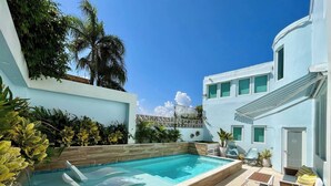 Una piscina con borde ondulado, rodeada de una terraza de madera y escalones que bajan al agua. En la terraza hay sillas de descanso y una pequeña mesa, y un edificio de fachada blanca y numerosas ventanas al fondo.