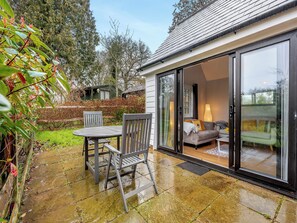 A patio with a table and chairs, a sliding glass door leading to a living area with a sofa and lamp, and a garden with trees and a fence. - 1 bedroom accommodation in Heathfield (Heathfield)