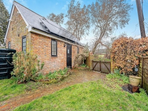 A brick house with a sloped roof, a wooden gate, and a garden with grass and plants. - 1 bedroom accommodation in Heathfield (Heathfield)