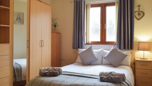 Ein Schlafzimmer mit Bett, Kissen, Nachttisch, Lampe, Schrank und einem Fenster mit Vorhängen.