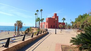 A coastal promenade with a stone walkway, palm trees, and a red building with a tower.