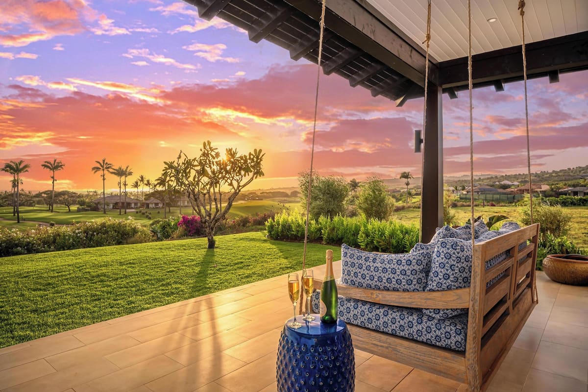 A patio with a swing, a table with a bottle and glasses, and a view of a golf course and sunset.