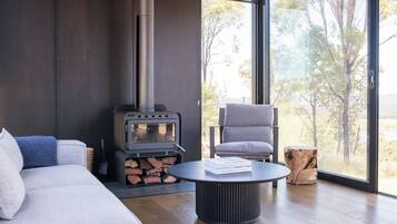 A modern living room with a sofa, a chair, a round table, a wood-burning stove, and a large window with a view of trees.