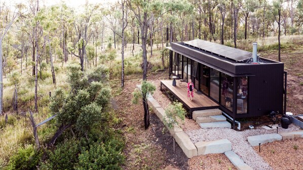 A modern, minimalist cabin with a deck and large windows, surrounded by a forested area.