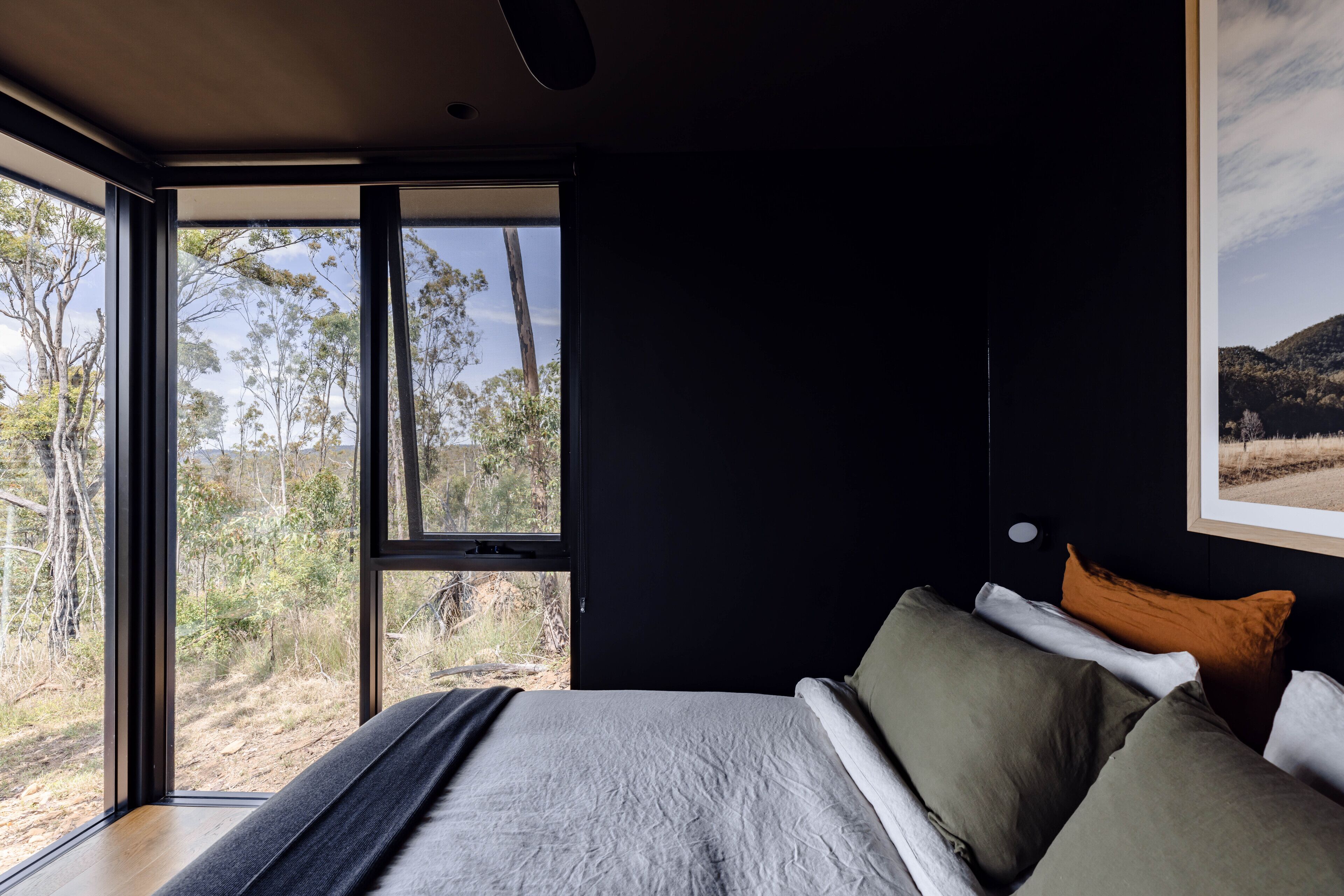 Een slaapkamer met een groot raam, een bed met grijze beddengoed, en een lijst met een afbeelding aan de muur.