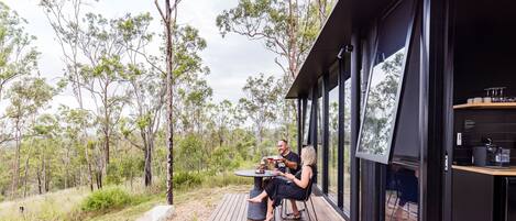 A modern outdoor dining area with a table and chairs, surrounded by a deck and large windows overlooking a forested landscape.