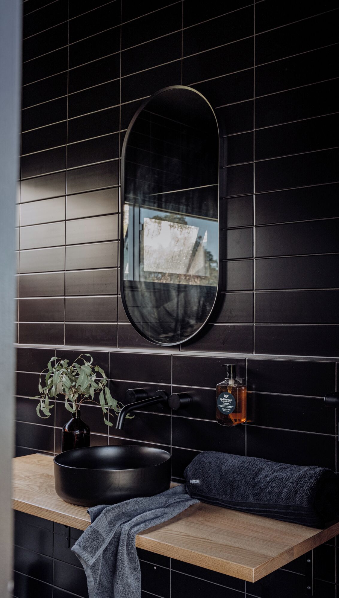 A bathroom with a black tiled wall, a round mirror, a sink with a black basin, a bottle of soap, and a towel on a wooden countertop.