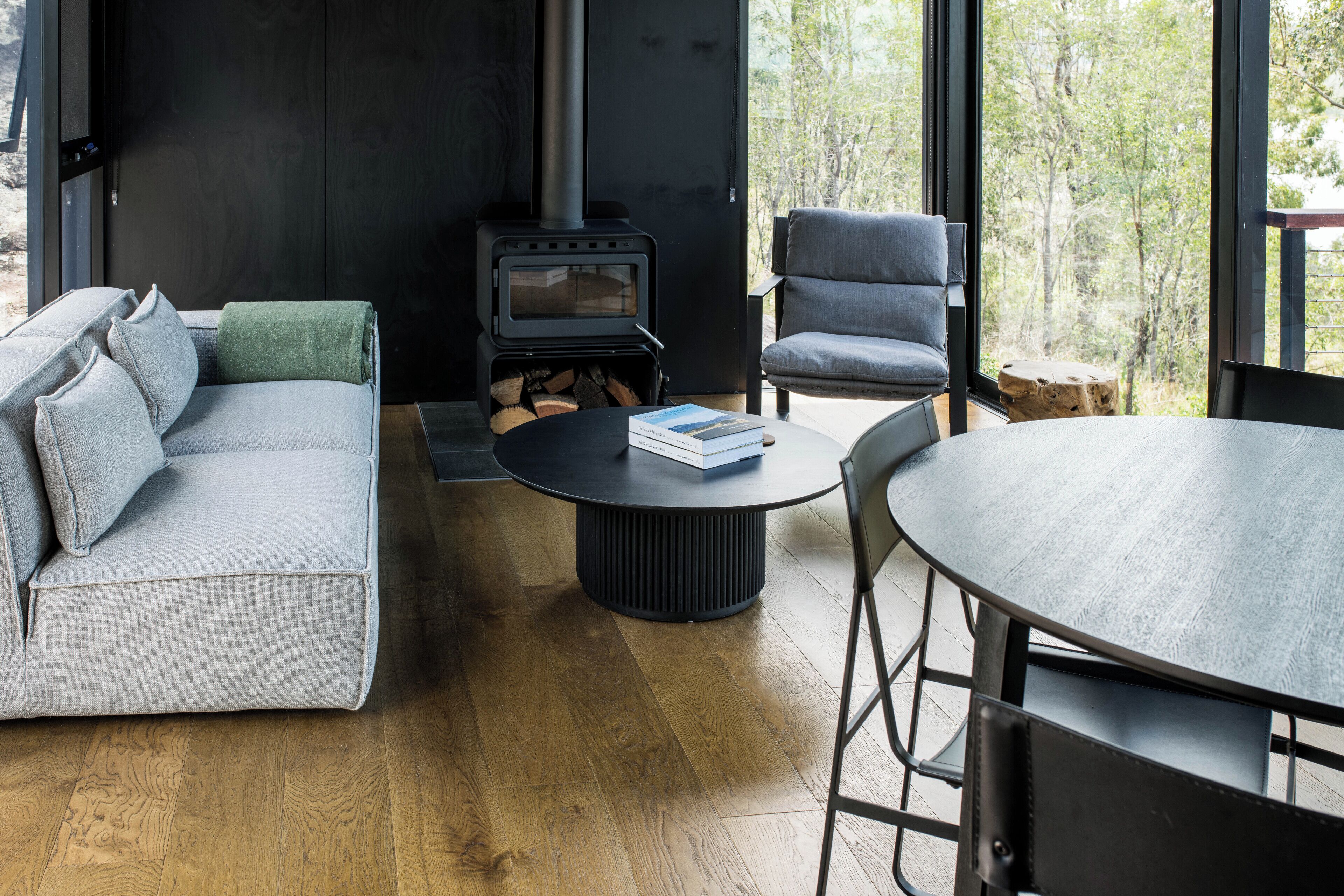 Una sala de estar moderna con sofá, sillón y mesa redonda, estufa de leña y vista a árboles a través de amplios ventanales.
