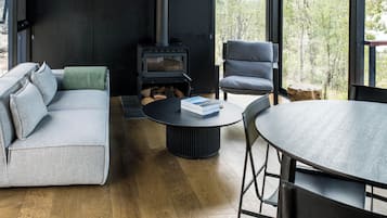 Una sala de estar moderna con sofá, sillón y mesa redonda, estufa de leña y vista a árboles a través de amplios ventanales.