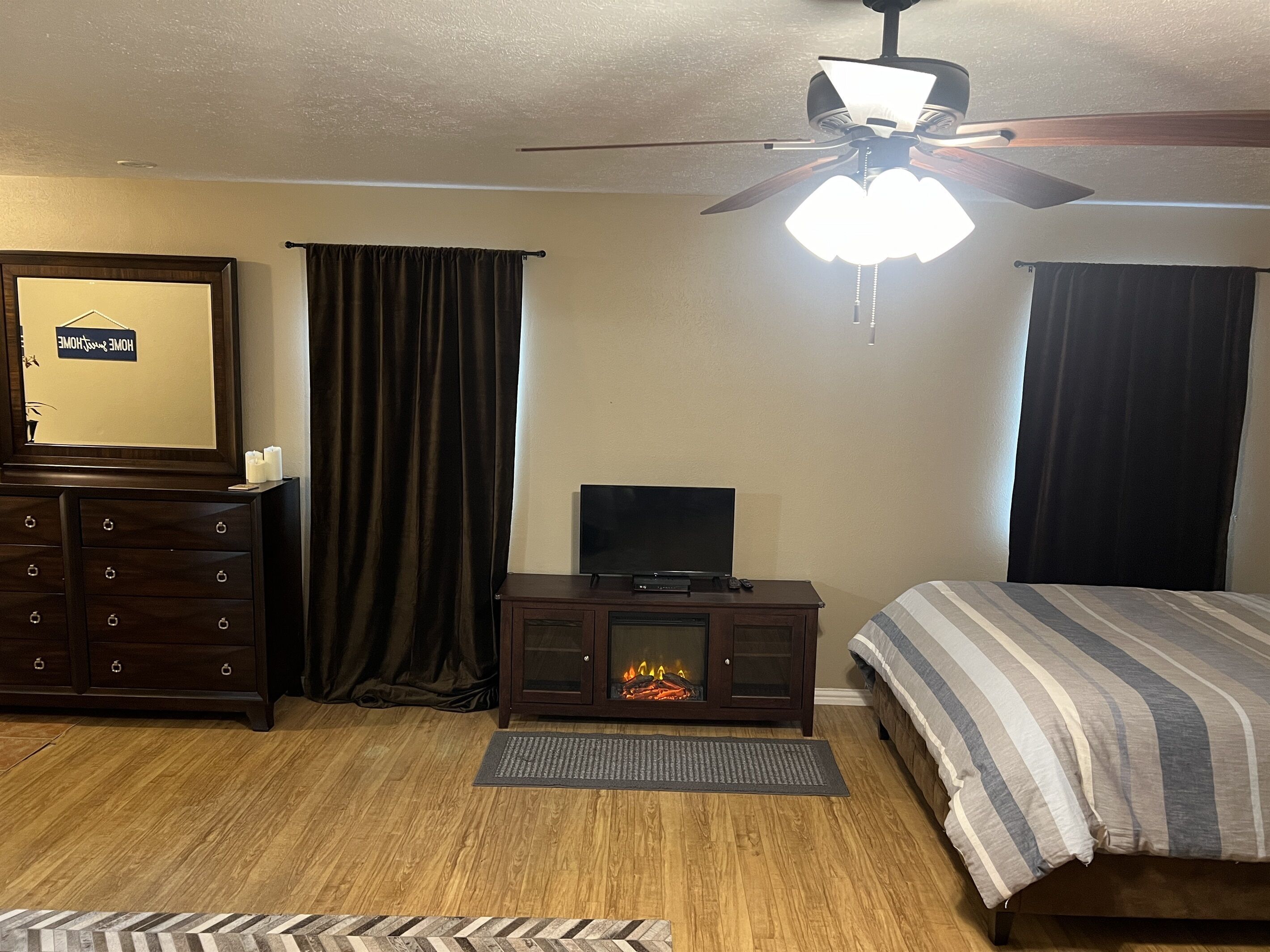 A bedroom with a bed, a TV, a fireplace, a ceiling fan, and a dresser.
