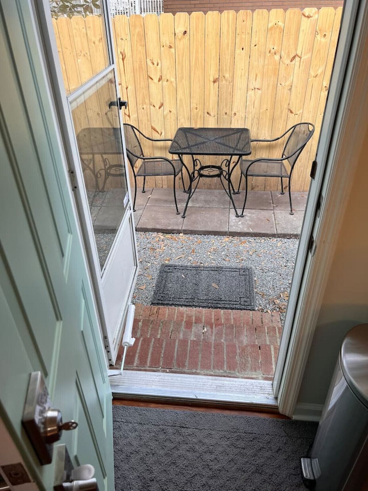 A view through a door into a patio area with a wooden fence, a metal table and chairs, and a doormat.