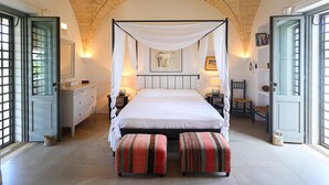 A bedroom with a four-poster bed, a white canopy, a wooden bench, a nightstand, a dresser, and a door with glass panels. - 3194 Masseria Il Carrubo by Perle di Puglia (Salve)