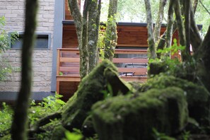Uma cabana rústica com parede de pedra e deck de madeira, rodeada por vegetação exuberante e árvores cobertas de musgo.