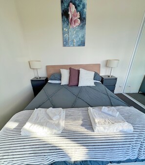 Een net opgemaakt bed met een gestreept laken, twee gevouwen handdoeken, een hoofdbord met een bloemschilderij en twee bedlampen op bijpassende nachtkastjes.